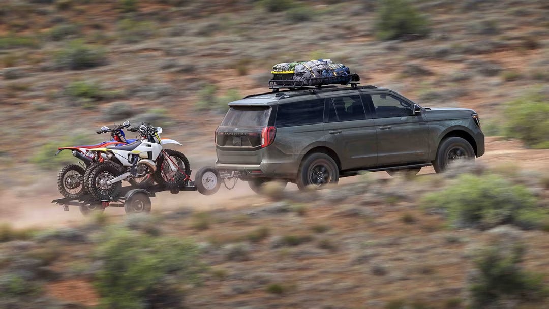 A green suv pulling a trailer with two dirt bikes on it.