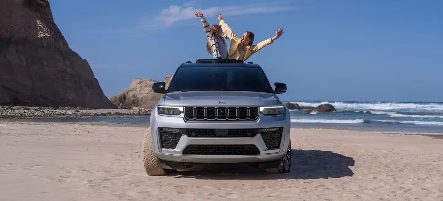 2026 Jeep Grand Cherokee parked on the beach with two people hanging out the sun roof.
